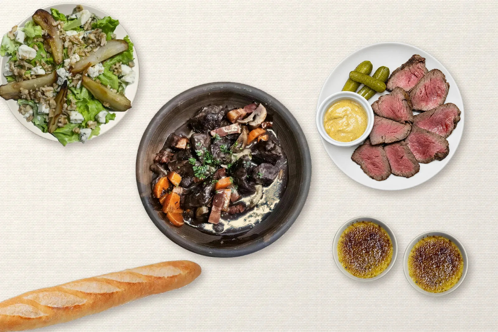 La mesa servida para dos sobre el mantel de Bocus, vista desde arriba: Boeuf bourguignon, Strogonoff, ensalada de pera y queso azul, Roast beef, baguette y crème brûlée.